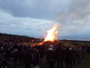 Många människor står runt en stor eld. Bilden är tagen i skymningen. I bakgrunden ser man havet